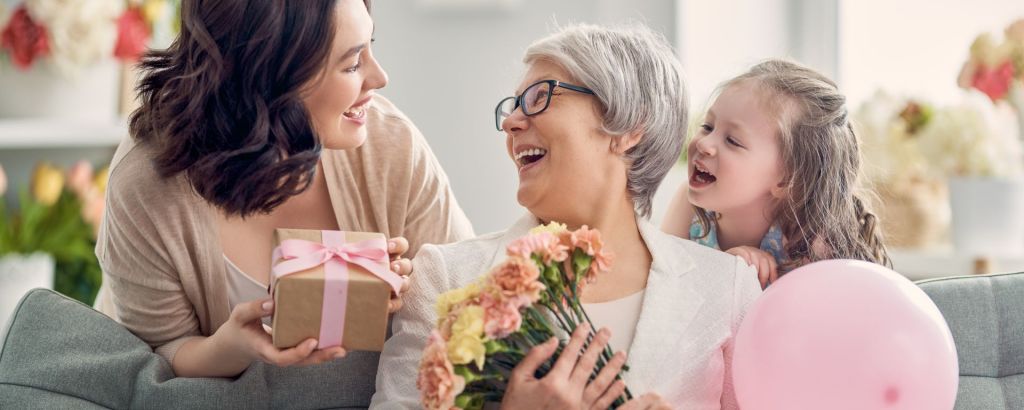 Child daughter is congratulating mom and granny giving them flowers and a gift. Grandma, mum, and girl smiling and hugging.